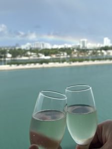 Image of an island in the background with two glasses of champagne clinking in the foreground from the cruise ship.